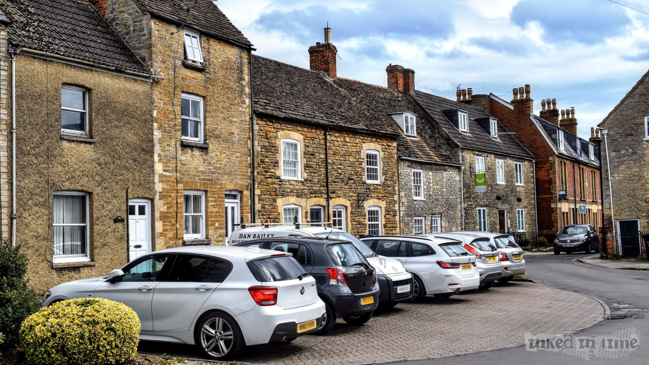 A view of the leftmost house in a terrace on Horsefair in Malmesbury, specifically 31 Horsefair. The house is part of a row of traditional stone and brick cottages with sloping roofs. The house at 31 Horsefair has a textured, rendered exterior in a muted tone, with two windows on the ground floor and a white front door. The neighboring houses display classic stonework and brick, with small windows and charming architectural details. Several cars are parked in front of the houses, including a white BMW and a black Vauxhall. The scene is calm, under an overcast sky, highlighting the historic charm of the area.