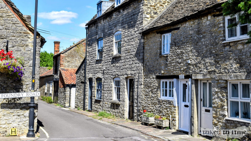 37 Horsefair, which is located at the edge of Horsefair near the entrance to Foundry Road in Malmesbury. The stone cottages lining the narrow street showcase traditional Cotswold architecture, with their rustic stone facades and charming details like small front gardens and hanging flower baskets. The road sign for Foundry Road is visible, adding to the quaint, picturesque feel of the area.