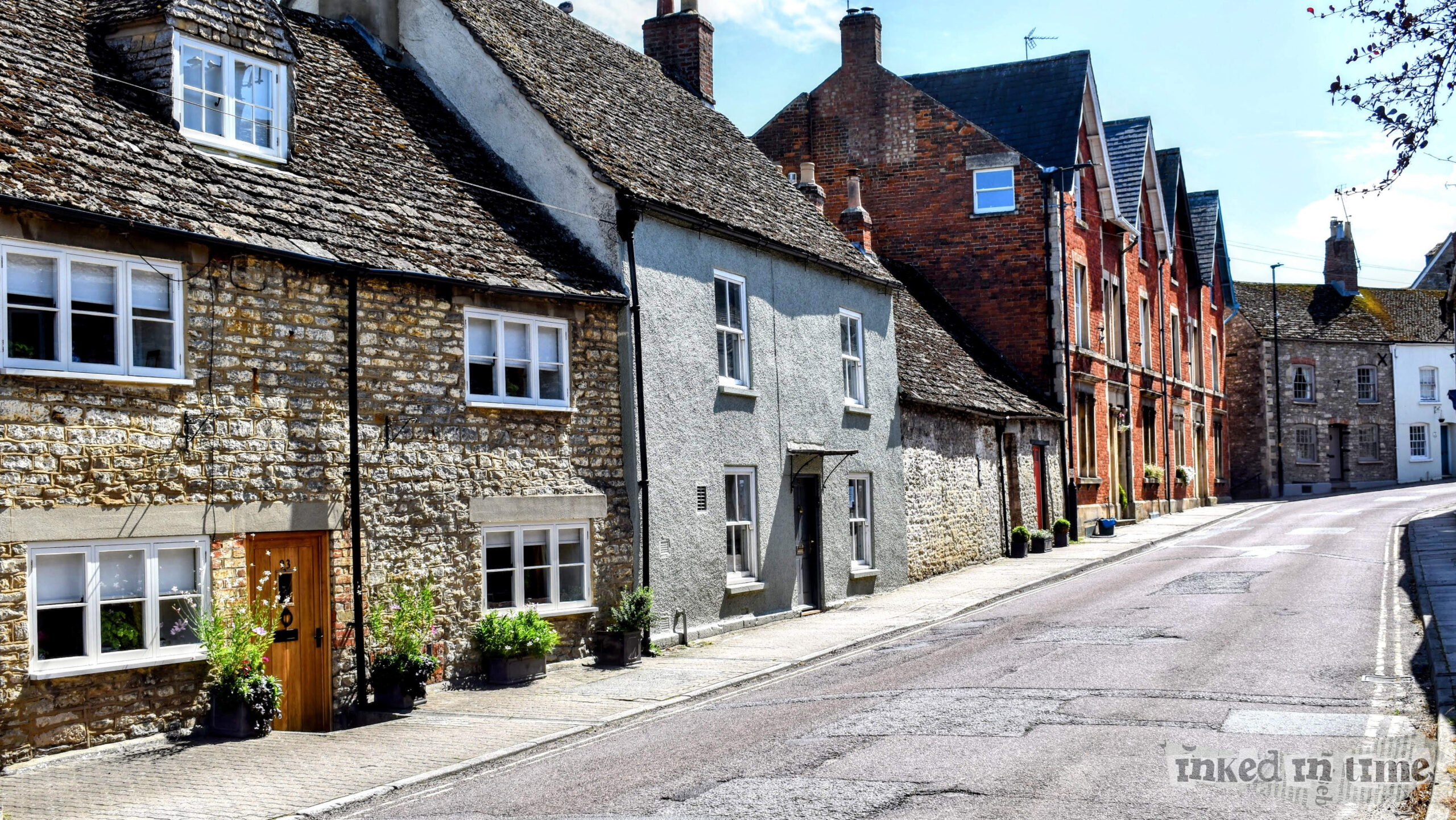 a view along Gloucester Road in Malmesbury, with 93 Gloucester Road in the foreground. The street scene highlights a mix of traditional stone and brick buildings, typical of the area, showcasing the historical architecture that Malmesbury is known for. The stone cottages in the foreground contrast with the red-brick buildings further down the street, giving a sense of the architectural variety found in this town. The well-maintained facades, flower pots, and the gently curving road add to the picturesque and serene atmosphere of this location.