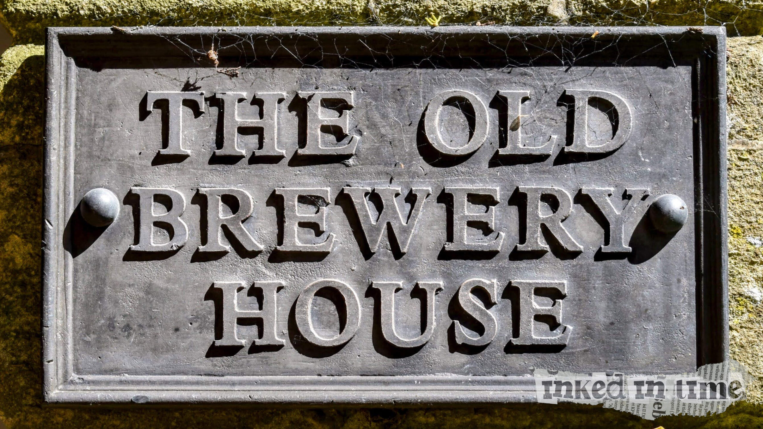 A close-up of a sign reading "THE OLD BREWERY HOUSE" in bold, capital letters. The sign appears to be made of metal with a weathered texture and is mounted on a stone wall. There are cobwebs visible in the corners of the sign, adding to its aged appearance.