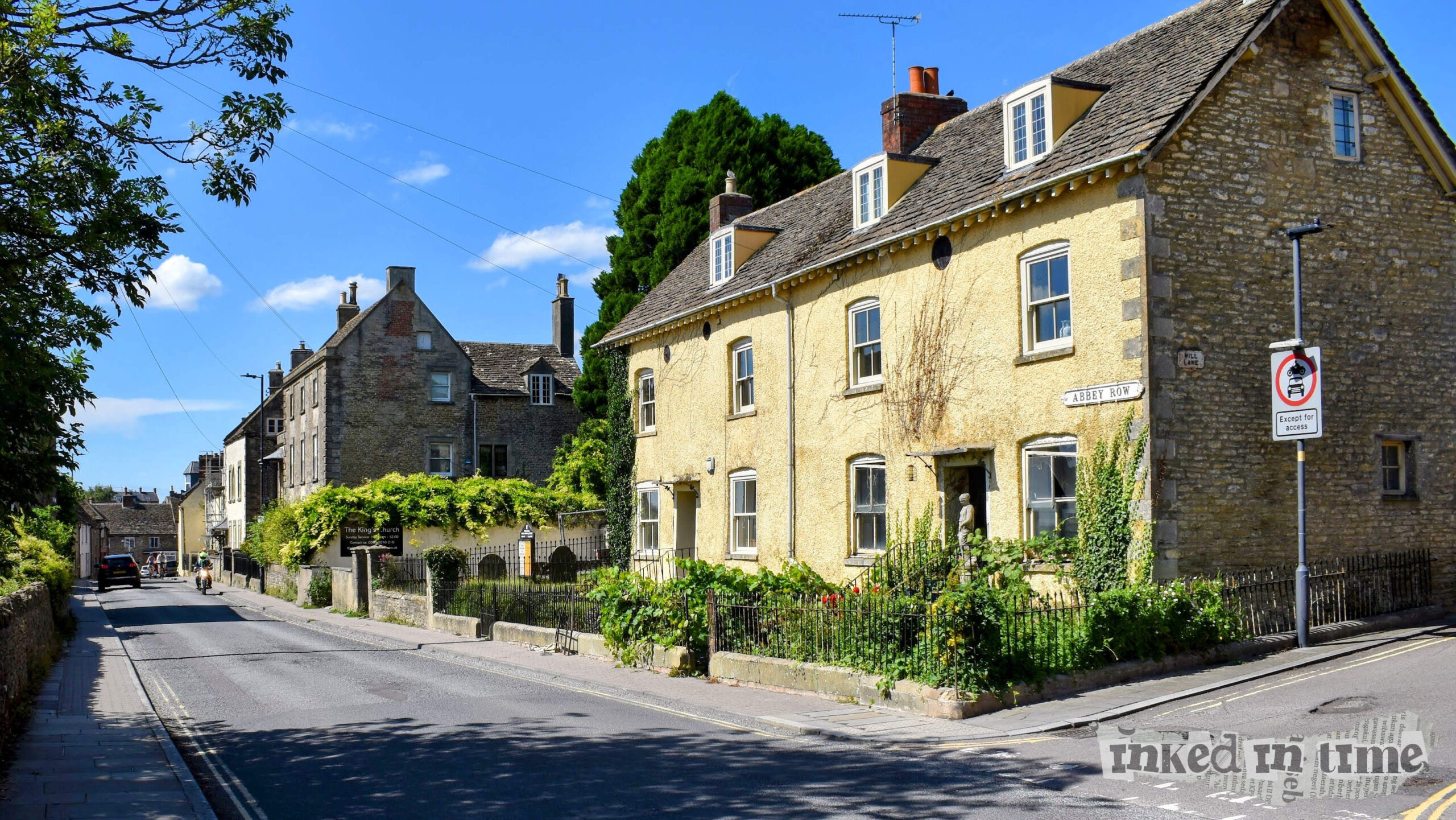 A scenic view of Abbey Row in Malmesbury, Wiltshire, with the perspective looking down the road toward the Triangle. The charming architecture, typical of English countryside towns, is evident in the stone buildings, with some covered in ivy and greenery. The blue sky and the peaceful atmosphere contribute to the overall picturesque setting.