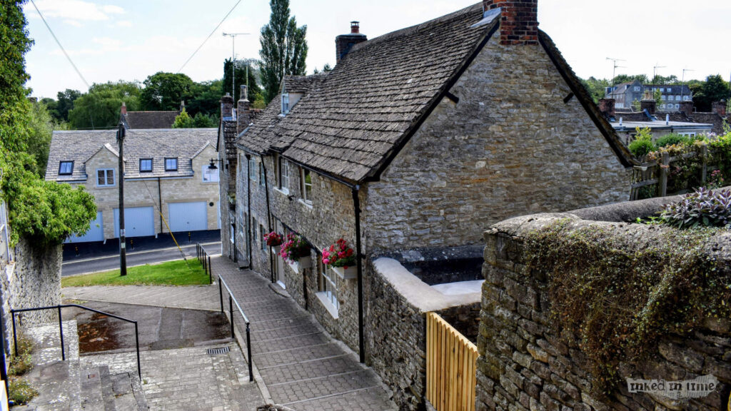 A picturesque view of Back Hill steps in Malmesbury, featuring a narrow, sloping path lined with traditional stone cottages. The cottages have steep, tiled roofs and small windows, some adorned with vibrant flower boxes. The stone-paved steps lead downhill, bordered by rustic stone walls. In the background, there are more modern buildings with garages, blending into the historic surroundings. The scene captures the charming, timeless character of Malmesbury, with its blend of old-world architecture and serene village atmosphere.