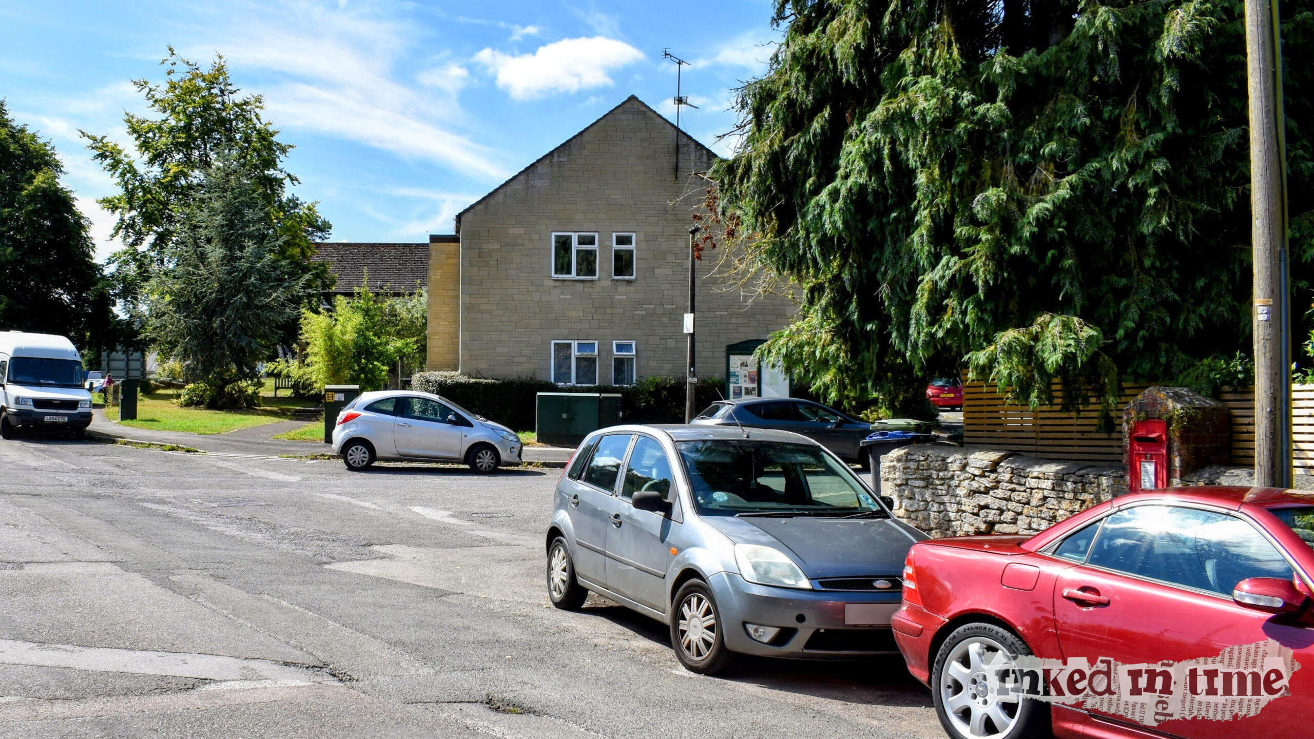 A quiet residential street with cars parked along the roadside and a large, leafy tree partially obscuring the view. The postbox is visible on the right-hand side, slightly obscured by a red car. The Black Horse was formerly located next to this postbox. A stone building and other houses can be seen in the background.