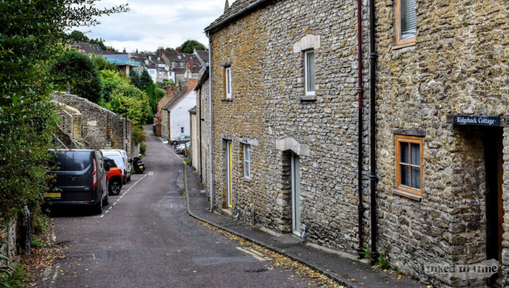 A view along Burnivale in Malmesbury, looking from the Bristol Street end towards the town. The narrow street is lined with historic stone cottages, including one with a sign reading "Ridgeback Cottage." The street is slightly sloped, with parked cars on the left side and a mix of greenery and stone walls on the right. The scene is tranquil, with overcast skies above, giving the area a peaceful and timeless atmosphere.