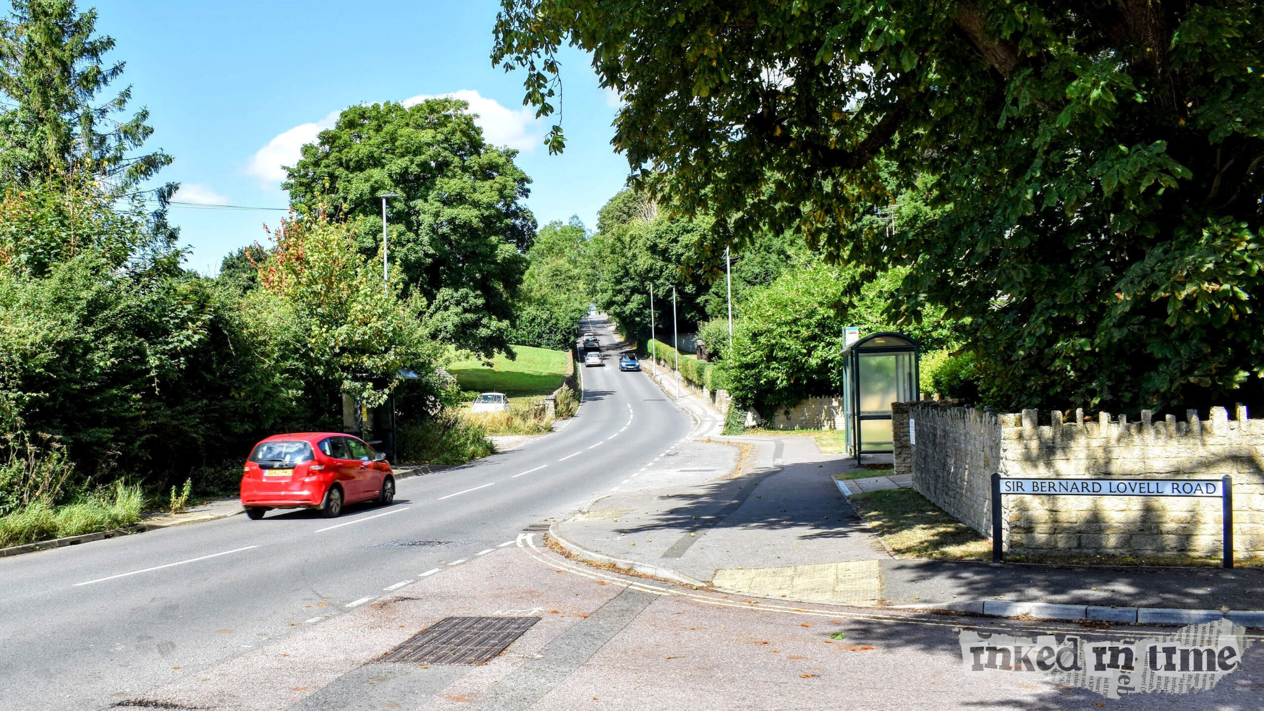 A view from the bottom of Cowbridge Hill looking towards Malmesbury. The road stretches ahead, lined with trees and greenery on both sides. A red car is driving along the road, and other vehicles can be seen in the distance. To the right, a stone wall with a sign that reads "Sir Bernard Lovell Road" is visible, along with a bus shelter and a paved sidewalk. The scene is framed by large trees, with clear skies above.