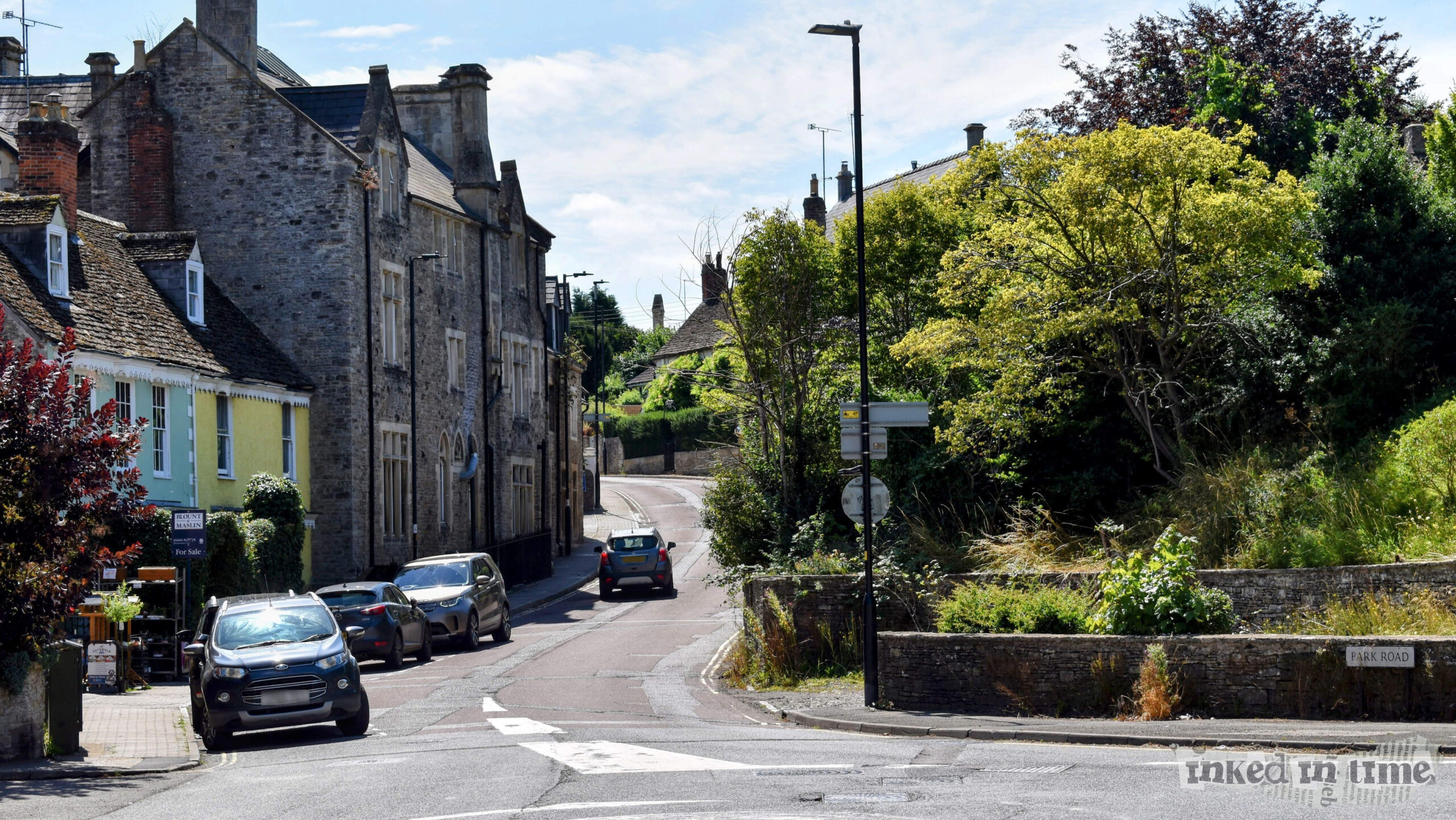 A view of Malmesbury looking up Gloucester Road with the entrance to Park Road on the right. The scene features historic stone buildings on the left, cars parked along the street, and lush greenery around the area. The road curves upwards into the distance.