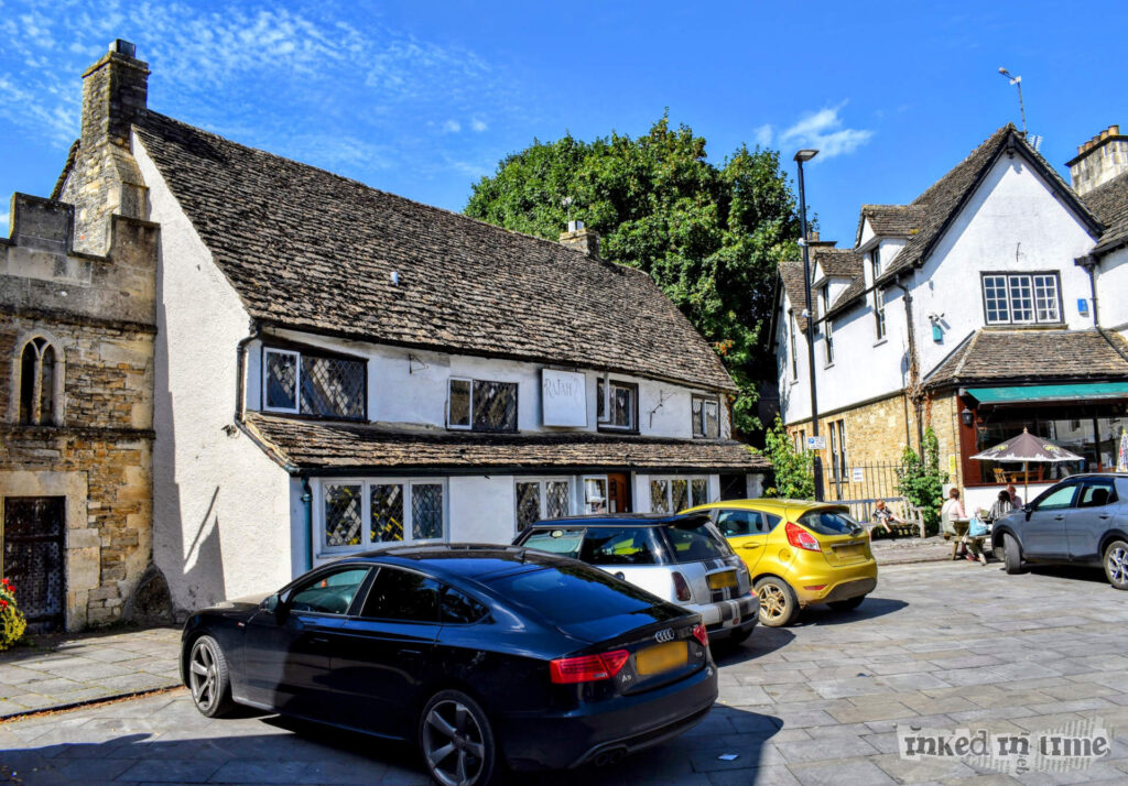 The image depicts the building that was formerly the Green Dragon near to Malmesbury's Market Cross. The focal point is a traditional stone building with a steep, slanted roof covered in weathered tiles. The building features small, leaded glass windows typical of older architecture. There is a sign above the door that indicates it is currently called the Rajah. Several modern cars are parked in front of the building on a cobblestone or paved area, providing a contrast between the old and new. In the background, there are other historic buildings, and a few people are sitting outside a cafe or pub, enjoying the day under a clear, blue sky.