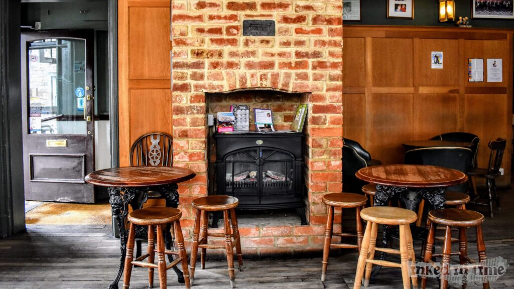 A cozy interior photograph taken inside The Whole Hog in Malmesbury. The focal point is a brick fireplace with a small black cast-iron stove inside it. Above the stove, a plaque with the word "JUBILEE" is embedded in the brickwork. In front of the fireplace, there are two round wooden tables surrounded by a few wooden stools with various designs. To the left, an open door provides a glimpse of the outside. The walls are paneled with wood, and there are framed pictures and notices hanging on them, contributing to the warm and inviting atmosphere of the pub.