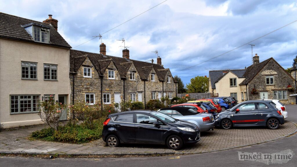 A view of the Horsefair in Malmesbury. The image shows a row of charming stone cottages with pitched roofs, nestled closely together. The cottages are surrounded by a small garden area with rose bushes and other plants. In the foreground, there is a parking area with several cars parked, including a black Ford Fiesta and a grey Fiat 500. The buildings are traditional in style, with stone walls and small windows, contributing to the historic and quaint atmosphere of the scene. The sky is overcast, adding to the tranquil and timeless feel of the location.