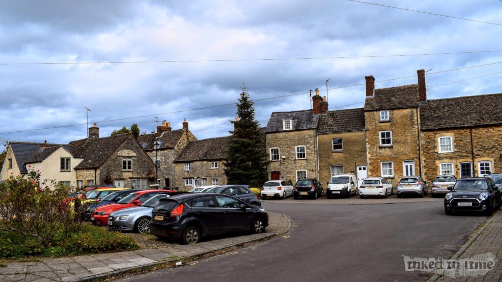 A view of the Horsefair in Malmesbury. The image shows a circular parking area surrounded by traditional stone cottages with pitched roofs. Several cars are parked around the perimeter, including a black Ford Fiesta and a white Mini Cooper. A large evergreen tree stands in the center of the image, adding greenery to the scene. The cottages have a mix of stone and brick facades, with some showing signs of weathering, giving them a historic and rustic appearance. The sky is overcast, contributing to the calm and serene atmosphere of this quaint town area.