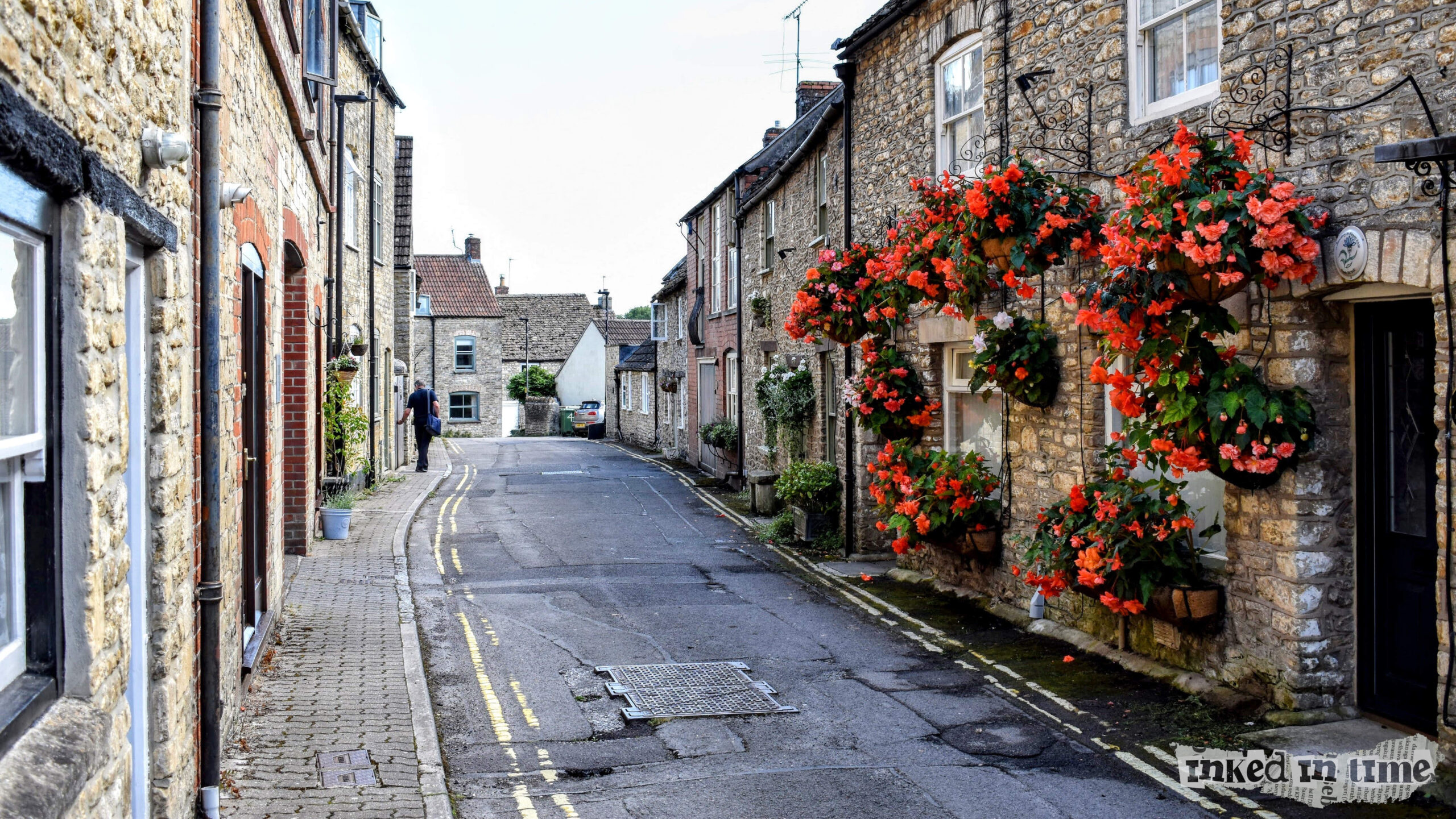 Ingram Street in Malmesbury, with a view toward Back Hill. The narrow, cobblestone street is lined with charming stone houses, many of which have vibrant hanging baskets overflowing with red and orange flowers. The old-world charm of the buildings, coupled with the winding road and quaint sidewalk, gives the street a cozy and picturesque feel. The person walking in the distance adds a touch of everyday life to this serene and beautifully maintained street.