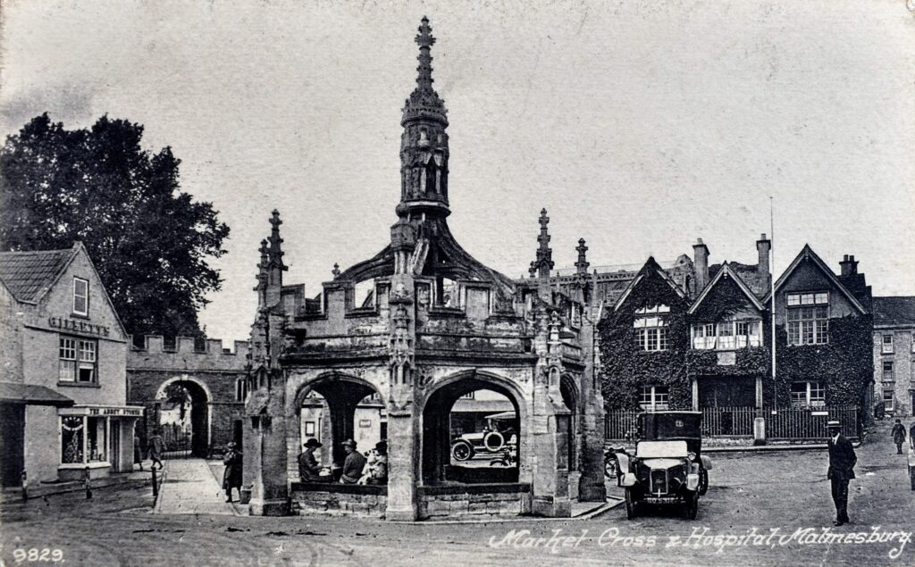 A black and white vintage postcard depicting the Market Cross in Malmesbury, with the Malmesbury Cottage Hospital visible in the background. The Market Cross is a medieval structure featuring a spire, arches, and intricate stonework. A few people are seated under the arches of the Market Cross, and early 20th-century automobiles are parked nearby. The surrounding buildings include a shop labeled "Gilbert's" on the left and "Abbey Stores" further down the street. The scene captures a glimpse of everyday life in Malmesbury during the early 1900s.