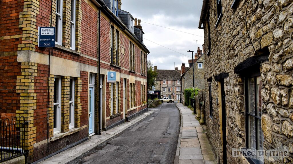 A photograph of a narrow street in Malmesbury, featuring Mansfield Terrace on the left side of the image. The red brick building of Mansfield Terrace has light yellow brick accents around the windows and doors, adding contrast to its facade. The street, St. Mary's Street, is lined with a stone pavement on the right side, where stone buildings with dark window frames and wooden doors are visible. The street leads toward a row of older, stone-built houses further down, with a car parked near the end of the street. The scene captures the historic and quaint character of Malmesbury.