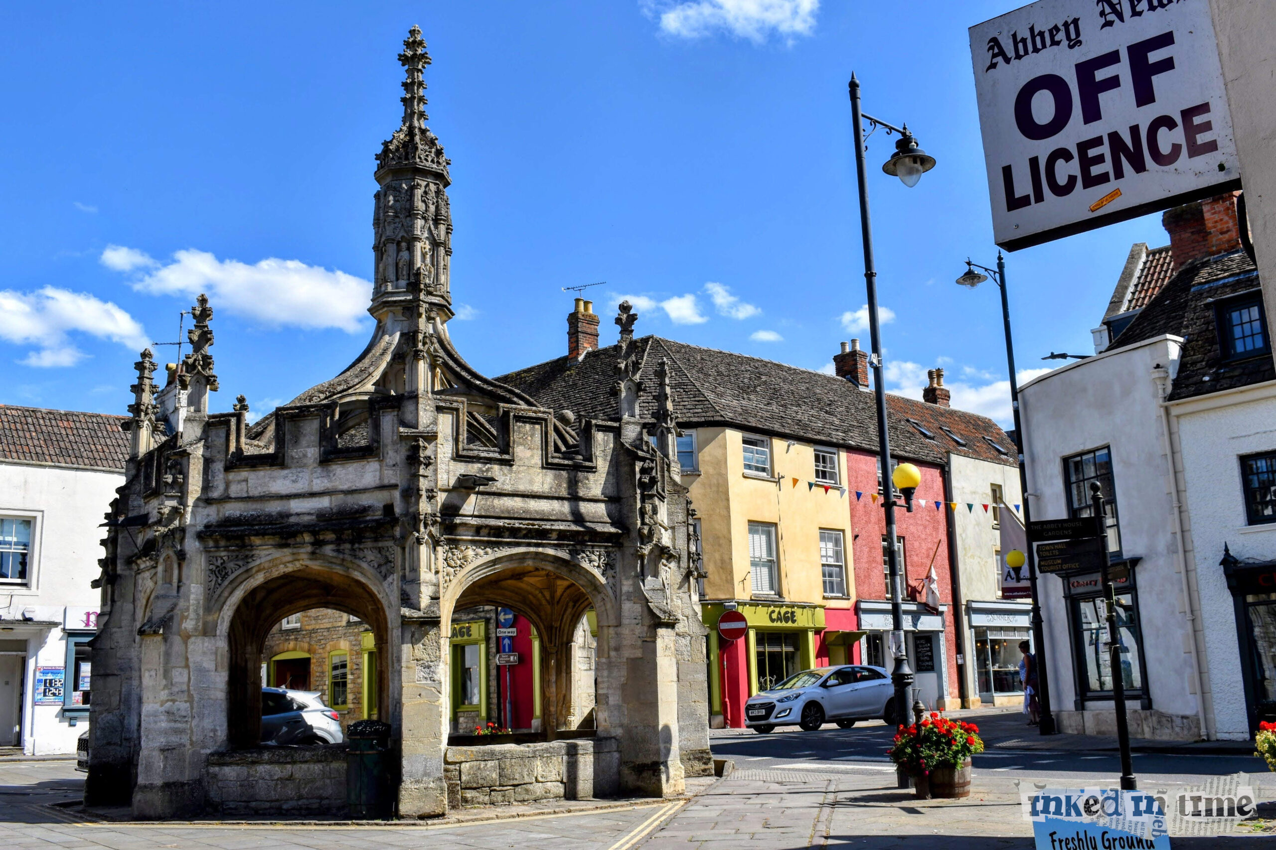 The Market Cross in Malmesbury, viewed from the entrance to Bird Cage Walk. The Market Cross is a historic and prominent feature in the town, reflecting the medieval architecture that is characteristic of many such structures across the UK. The scene includes colorful buildings in the background, with a clear blue sky adding contrast to the stone structure.