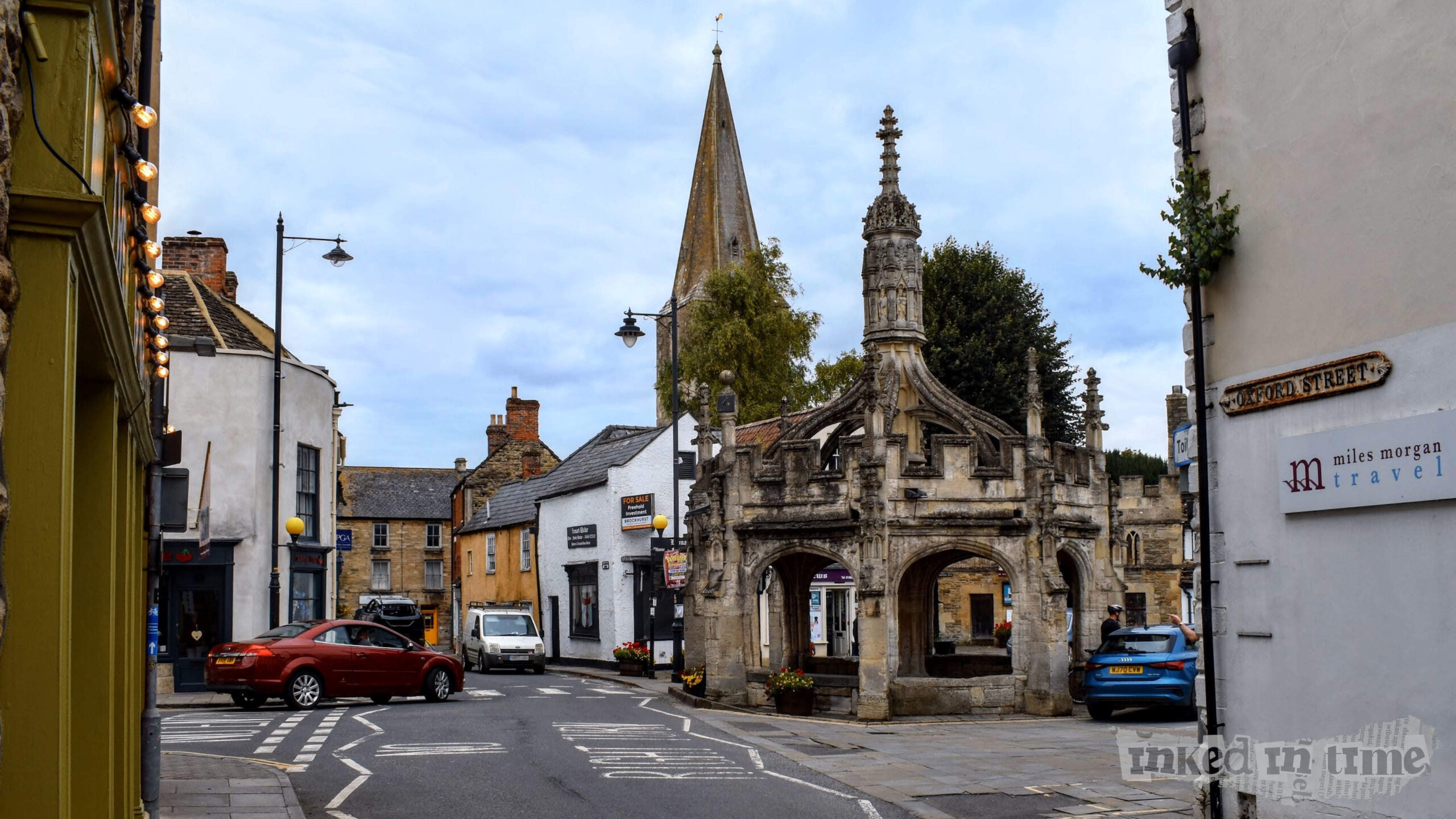 A view of the Market Cross in Malmesbury, seen from Oxford Street. The historic stone structure of the Market Cross stands prominently on the right, with its intricate Gothic design and tall spire visible against the overcast sky. In the background, the spire of a church rises above the surrounding buildings, adding to the town's historic charm. The street scene includes a few cars, a red one turning the corner and a blue one parked near the Market Cross. On the left, a building with green paint and string lights adds a touch of modernity to the otherwise historic setting. The scene is peaceful, capturing the blend of old and new in this quaint town.