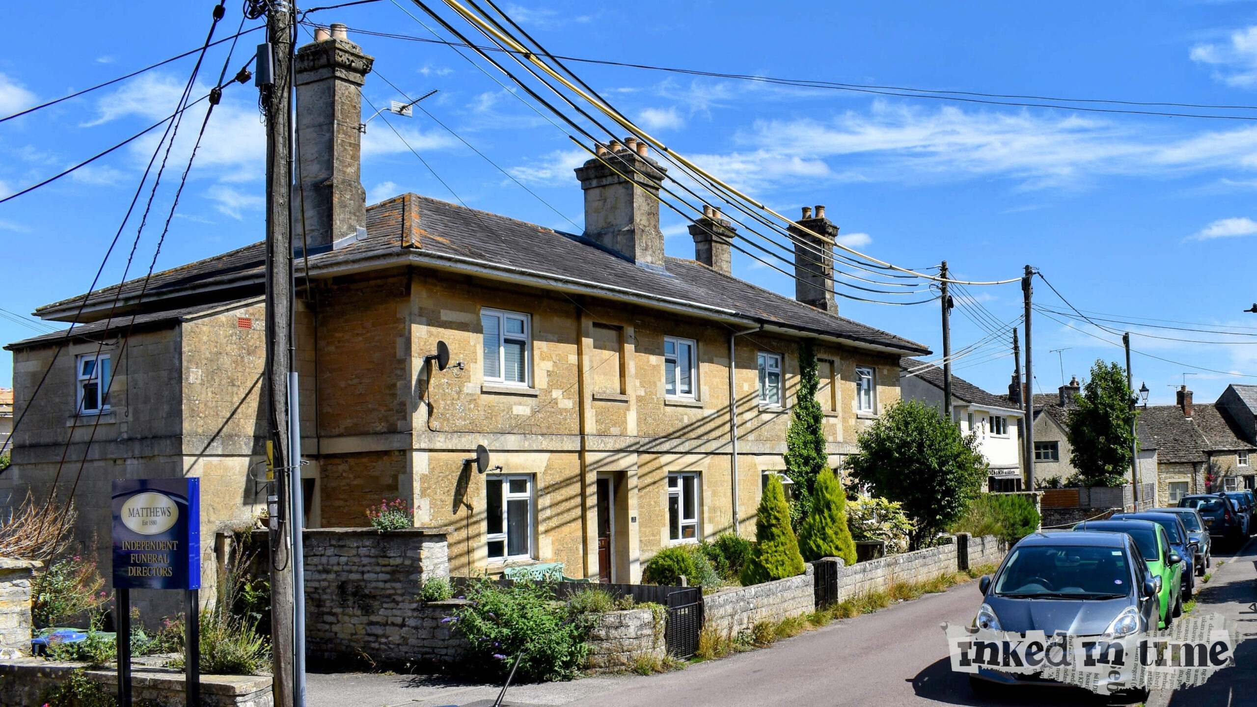 A row of stone cottages under a clear blue sky, with power lines overhead and cars parked along the narrow street. The cottage at the furthest end of the row was formerly Malmesbury police station. A sign for "Matthews Independent Funeral Directors" is visible in the foreground.