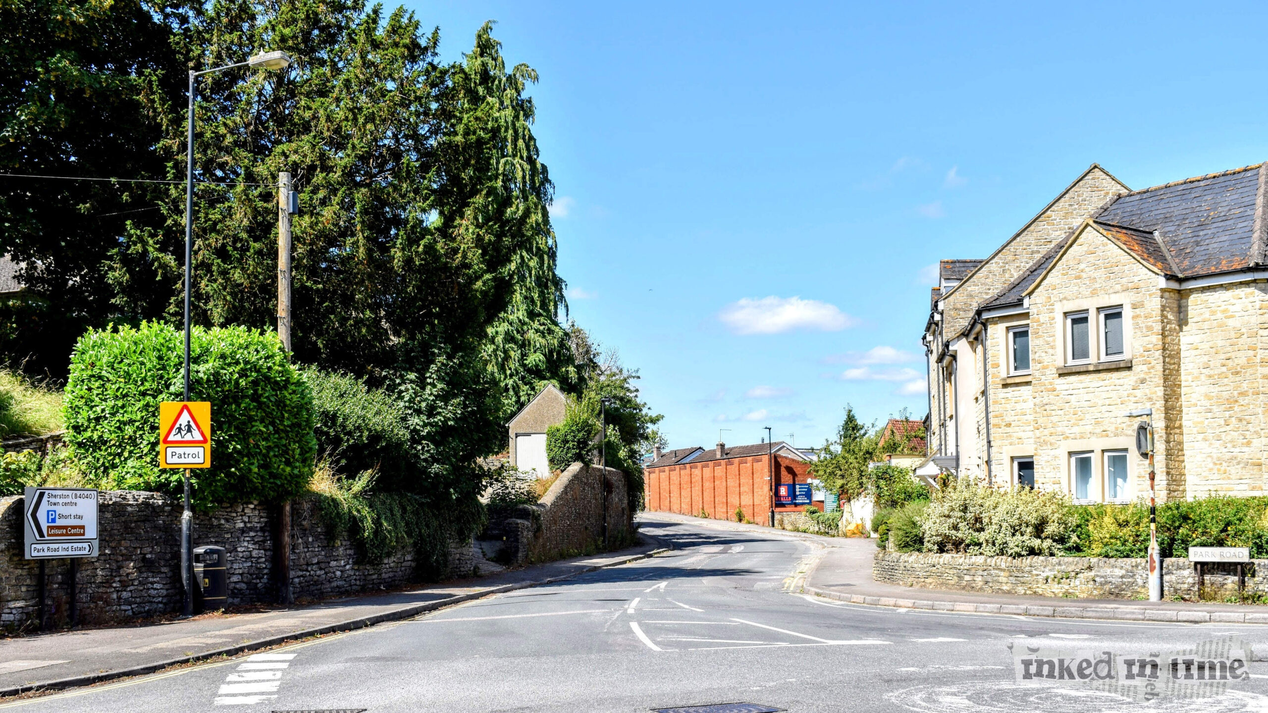 A view down Park Road from the bottom of Gloucester Road, featuring a road curving gently to the right. On the left side, there is dense greenery with a "Patrol" sign visible, while on the right, a stone building stands near the intersection. In the distance, a brick building is visible, with a clear blue sky above.