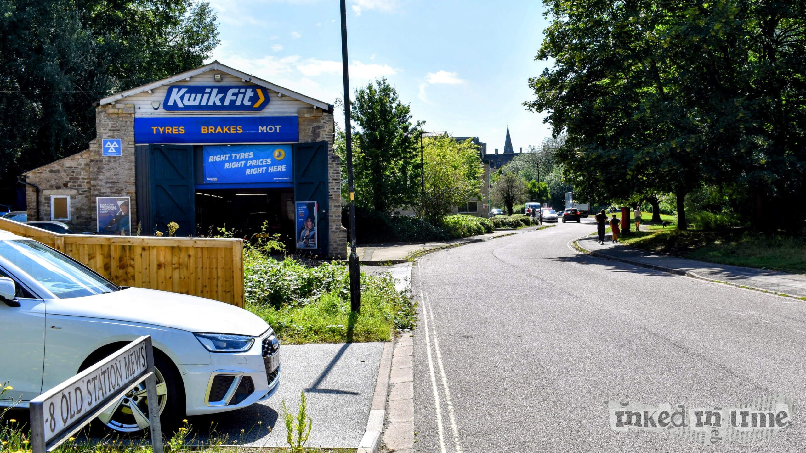 A sunny street scene featuring a Kwik Fit garage on the left side, housed in the only remaining building from Malmesbury's historic railway station. The garage displays a blue and white sign that reads "Tyres, Brakes, MOT." A white car is partially visible in the foreground next to a sign that says "8 Old Station Mews." The street curves to the right, lined with trees and a few people walking on the sidewalk. The area appears to be a quiet, residential neighborhood.