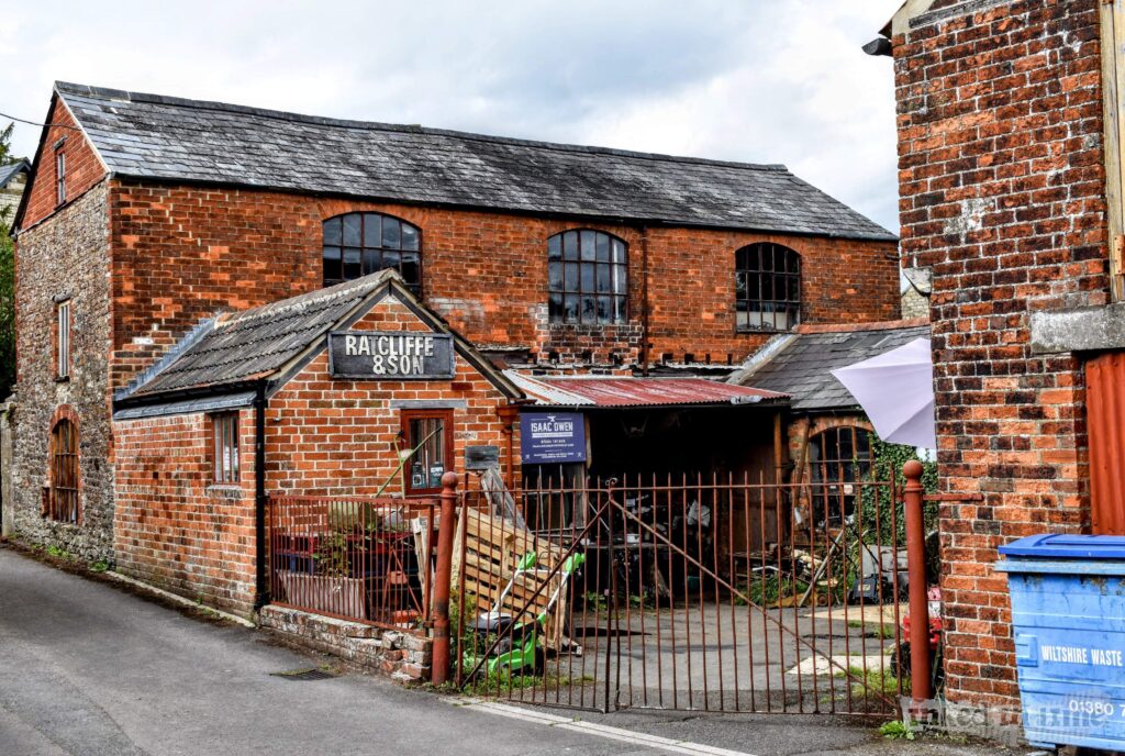 A view of Ratcliffe and Son on Foundry Road in Malmesbury. The building is an old, weathered brick structure with large arched windows and a slate roof, indicative of its industrial past. The main entrance is marked by a sign that reads "Ratcliffe & Son" above a small porch. The building has an aged, rustic appearance, with some areas showing signs of wear. A metal gate surrounds the entrance, and various tools and materials are visible inside the open area.