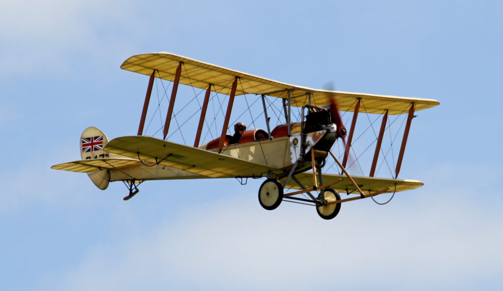 a Royal Aircraft Factory B.E.2, a British single-engine biplane used during World War I. The aircraft has a distinctive design featuring two open cockpits arranged in tandem, one behind the other, with the pilot typically sitting in the rear seat and the observer or instructor in the front. The plane has a fabric-covered frame with a prominent upper and lower wing structure, connected by multiple struts and wires, characteristic of early biplane designs. The engine is mounted at the front, driving a two-blade wooden propeller. The aircraft bears British military markings on its tail, with the Union Jack visible. The overall design reflects the rudimentary and exposed nature of early military aviation.