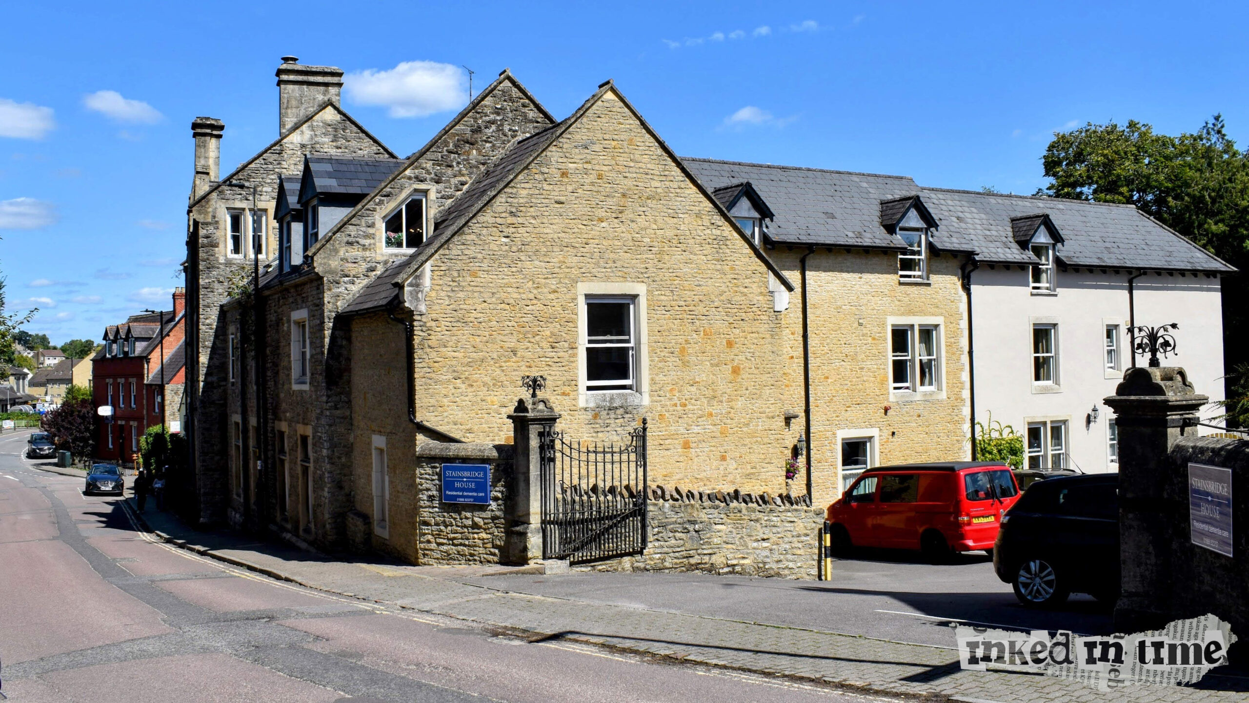 This image captures Stainsbridge House in Malmesbury, with the view directed toward the roundabout and the Co-op supermarket. The architectural style of Stainsbridge House is in keeping with the traditional stone construction seen throughout the town, with distinctive dormer windows and a gated entrance visible in the foreground. The road curves gently, leading down toward other buildings in the distance, including a striking red-bricked structure.