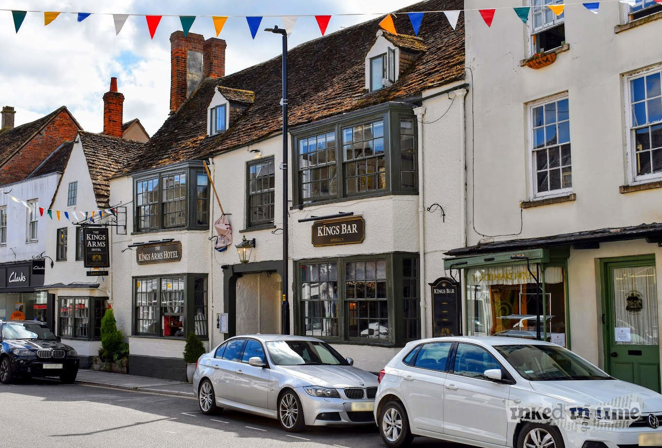 A view of Malmesbury High Street, featuring a historic row of buildings with a traditional English aesthetic. The central focus is on "The Kings Arms Hotel" and "Kings Bar," housed in an old, two-story building with a sloping, weathered roof and dormer windows. The facade is painted in light colors, with dark trim around the windows and doors, giving it a classic look. The windows are large with multiple panes, and a sign hangs above the entrance. Nearby shops, including a Clarks store, can be seen, and cars are parked along the street. Colorful bunting flags are strung across the street, adding to the festive and welcoming atmosphere. The sky is partly cloudy, with patches of blue visible, contributing to the bright and lively scene.