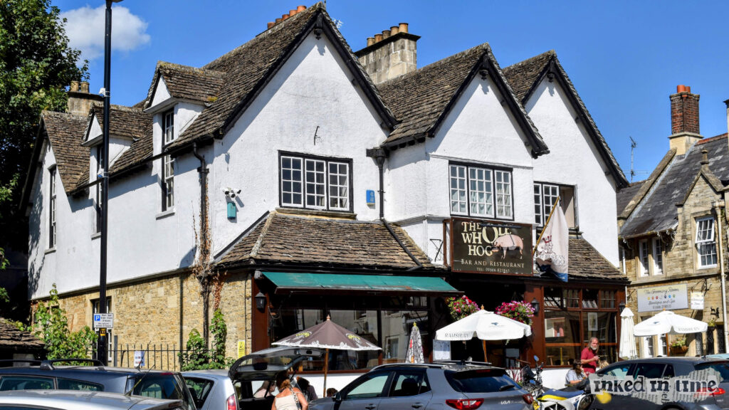 "The Whole Hog," a bar and restaurant in Malmesbury. Interestingly, this building has a historical significance, as it served as Malmesbury Cottage Hospital from late 1897 until the end of 1926. The building retains its traditional architectural charm, with steeply pitched roofs and whitewashed walls, characteristic of the local style. The exterior now includes signage for the restaurant and an outdoor seating area, reflecting its modern-day function as a dining establishment.