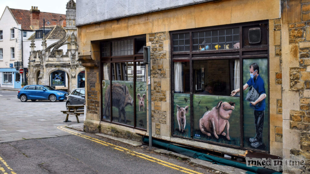 A view of the side of The Whole Hog in Malmesbury. The image features large windows with painted murals on them, depicting pigs and a man feeding them from a container labeled "Dreams." The artwork is vibrant and adds a creative touch to the building's stone facade. In the background, the historic Malmesbury Market Cross is visible, adding a sense of place and history to the scene. The area is paved, with a picnic bench outside and a couple of cars parked nearby. The overall atmosphere is quaint and lively, reflecting the town's blend of tradition and modern artistic expression.