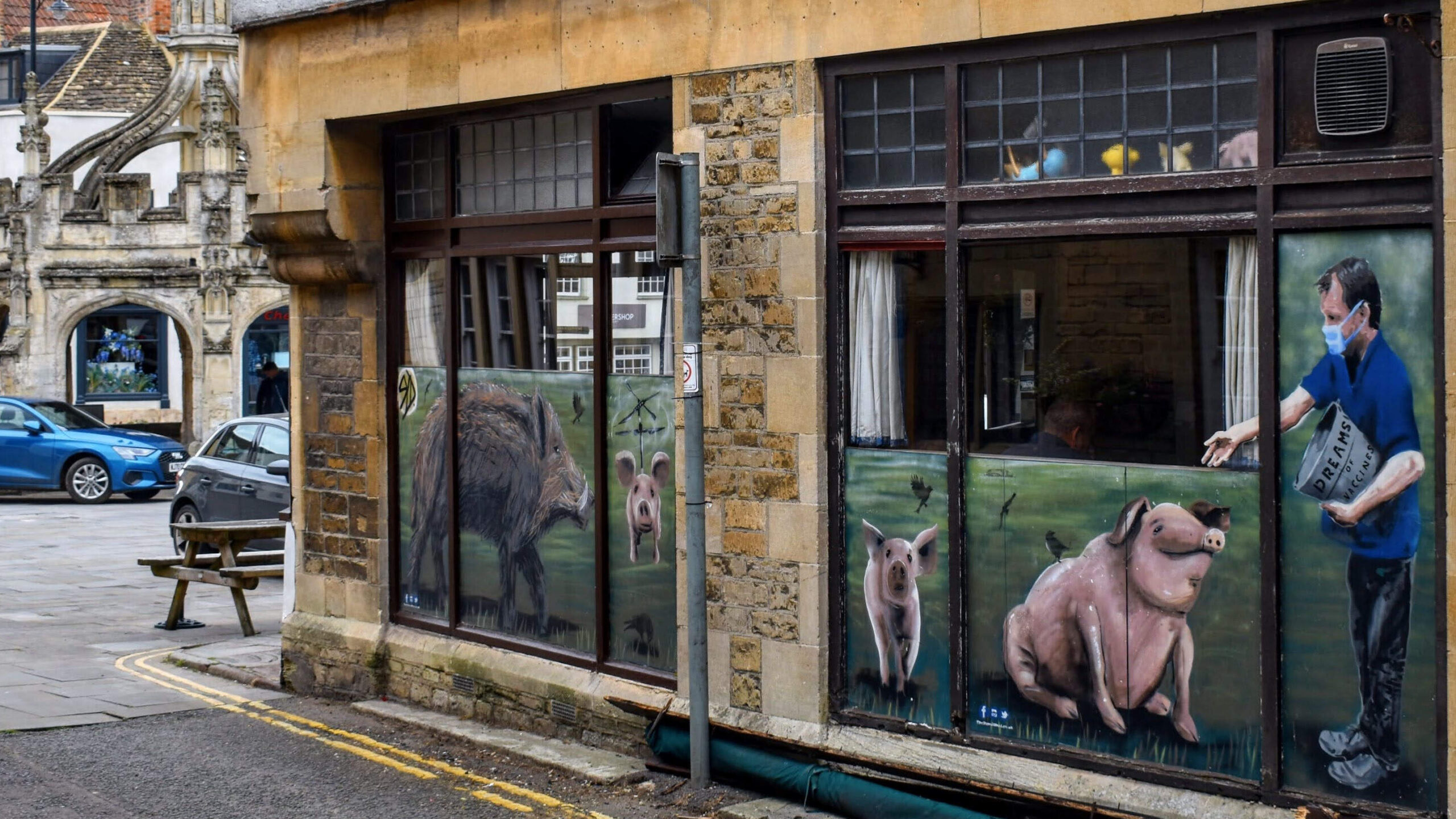 A view of the side of The Whole Hog in Malmesbury. The image features large windows with painted murals on them, depicting pigs and a man feeding them from a container labeled "Dreams." The artwork is vibrant and adds a creative touch to the building's stone facade. In the background, the historic Malmesbury Market Cross is visible, adding a sense of place and history to the scene. The area is paved, with a picnic bench outside and a couple of cars parked nearby. The overall atmosphere is quaint and lively, reflecting the town's blend of tradition and modern artistic expression.