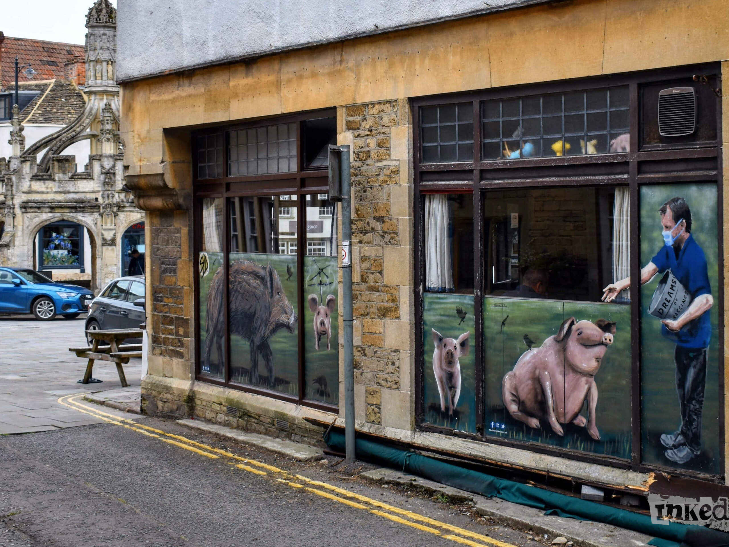 A view of the side of The Whole Hog in Malmesbury. The image features large windows with painted murals on them, depicting pigs and a man feeding them from a container labeled "Dreams." The artwork is vibrant and adds a creative touch to the building's stone facade. In the background, the historic Malmesbury Market Cross is visible, adding a sense of place and history to the scene. The area is paved, with a picnic bench outside and a couple of cars parked nearby. The overall atmosphere is quaint and lively, reflecting the town's blend of tradition and modern artistic expression.