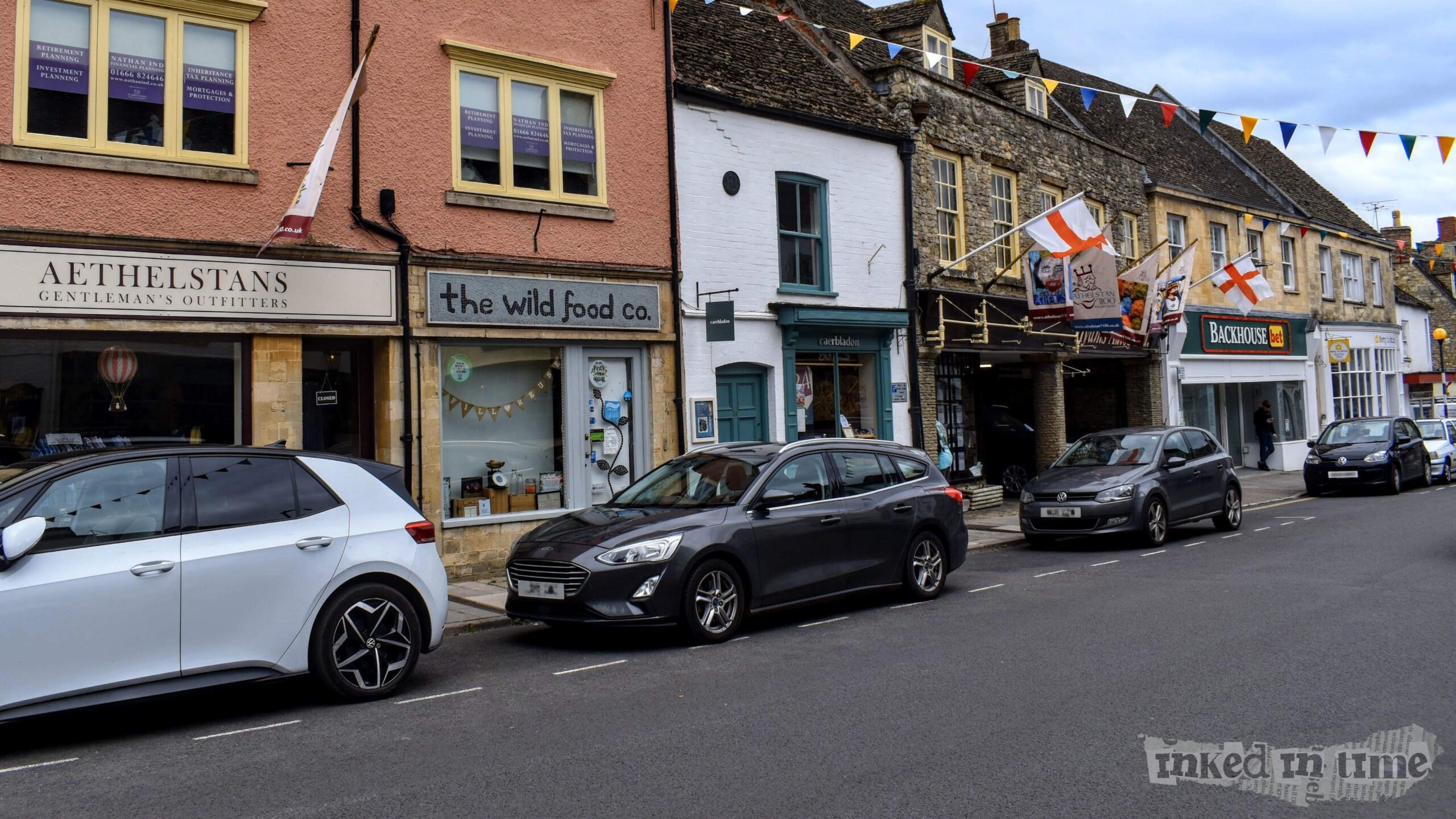 A view of The Wild Food Co in Malmesbury, located in a row of historic buildings on a bustling street. The shop has a stone facade with large windows displaying various products, and a sign above that reads "the wild food co." The building next door is painted white with a teal-colored door and window frames, labeled as "cartablanco." On the left, there is Aethelstans Gentleman's Outfitters with a peach-colored exterior. Several cars are parked along the street, and colorful bunting is strung across the road, adding a festive touch to the scene. In the past, this location was once home to Mr. Pitt's china shop, reflecting the evolving nature of the town's businesses. The overall atmosphere is quaint and lively, with a mix of tradition and modernity.