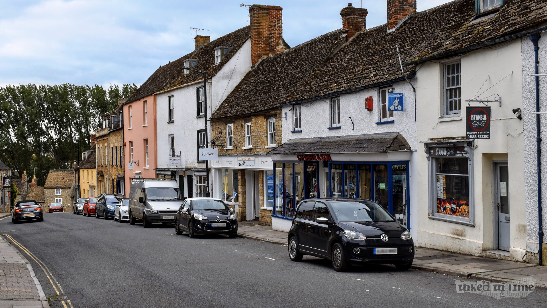 A view looking down the town hill in Malmesbury. The street is lined with historic buildings featuring a mix of stone and painted facades, including shops and businesses such as "Istanbul Grill" and "Strakers." The road is slightly sloped, with cars parked along the left side. The buildings have charming, traditional architecture, including sloping roofs and chimneys. The scene is peaceful, with overcast skies and trees visible in the background, contributing to the small-town charm of the area.