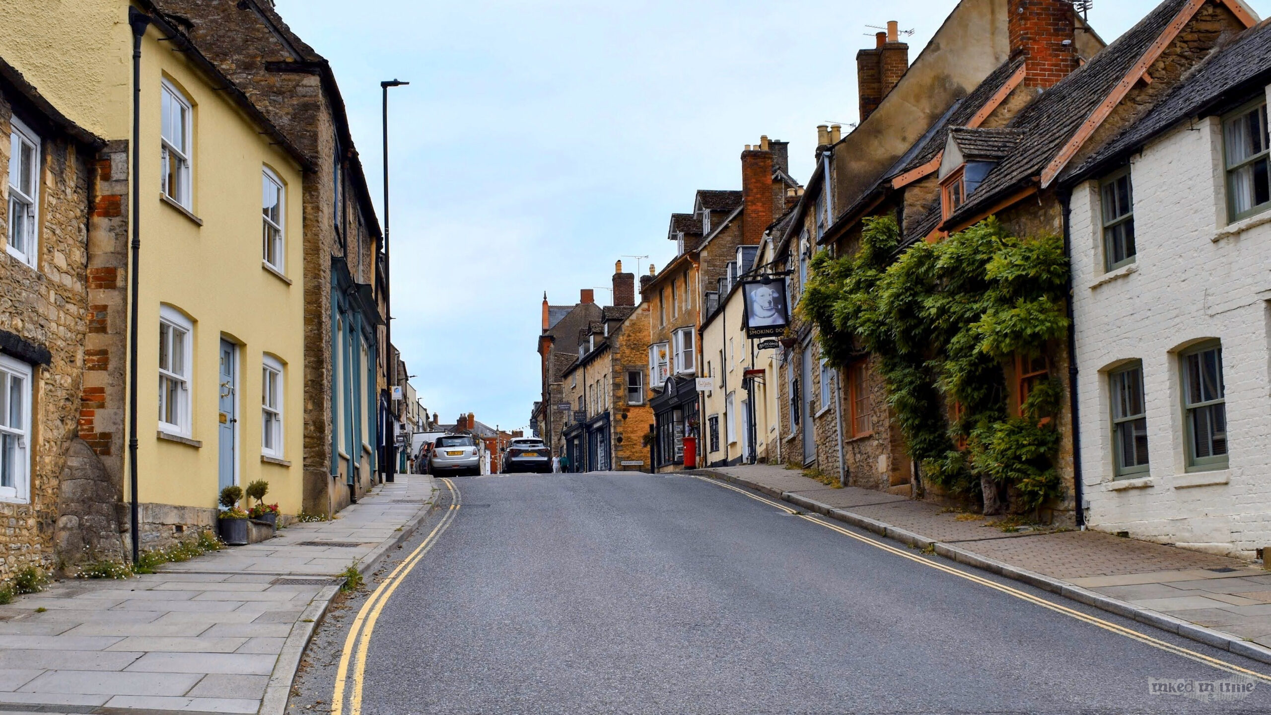 A view looking up the town hill in Malmesbury. The street is flanked by historic buildings with a mix of stone, brick, and painted facades, including some covered in greenery. The road slopes upwards, with a narrow sidewalk on the left and right. A pub sign with a classic design hangs on one of the buildings on the right side of the street. The scene captures the quaint and charming character of the town, with traditional architecture and a peaceful atmosphere under an overcast sky.