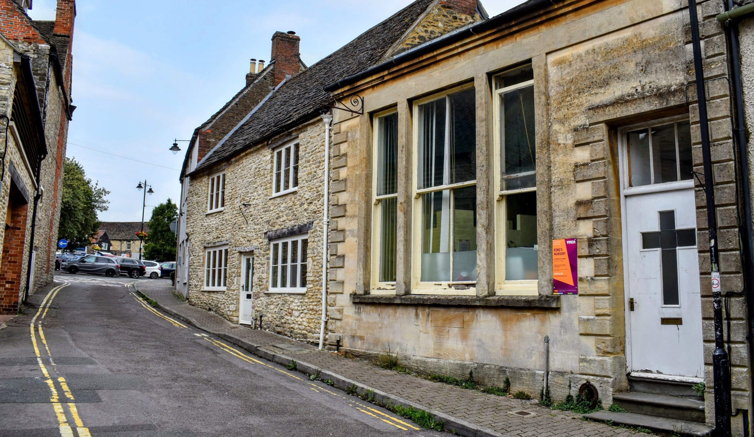 A photograph taken on Silver Street in Malmesbury, featuring the YMCA Hall in the foreground on the right side. The YMCA Hall is a stone building with large windows and a white door, which has a cross design on it. A sign for "King's Nursery" is posted on the wall next to the door. The street is narrow with a slight incline, leading up to a small intersection where a few cars are parked. The buildings along the street are constructed from traditional stone, typical of the area, and a lamppost is visible further up the road. The scene captures a quiet moment on a historic street in Malmesbury.