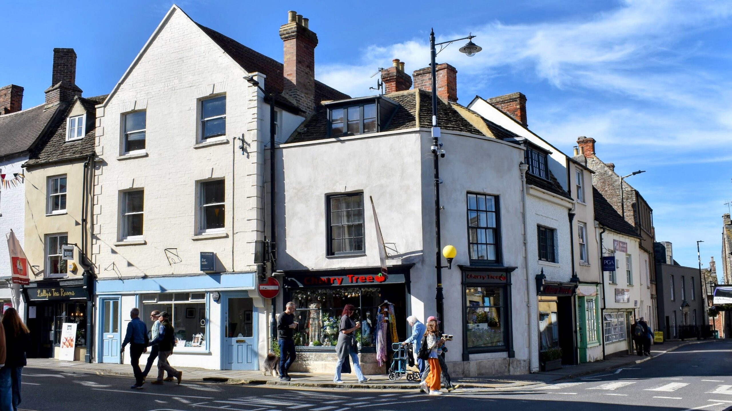 A sunny view of a bustling street in Malmesbury featuring traditional multi-story buildings. The Cherry Tree shop, with a sign prominently displayed, is located at 1 High Street. Pedestrians and a couple with a dog are walking along the pavement, passing various shops, including Lally's Tea Rooms.