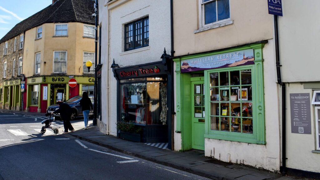 Street-level view of Gloucester Street in Malmesbury, showcasing The Cherry Tree shop front with a dark grey façade and a red sign, next to Wheeler’s Retreat Cycling Emporium with a green front.